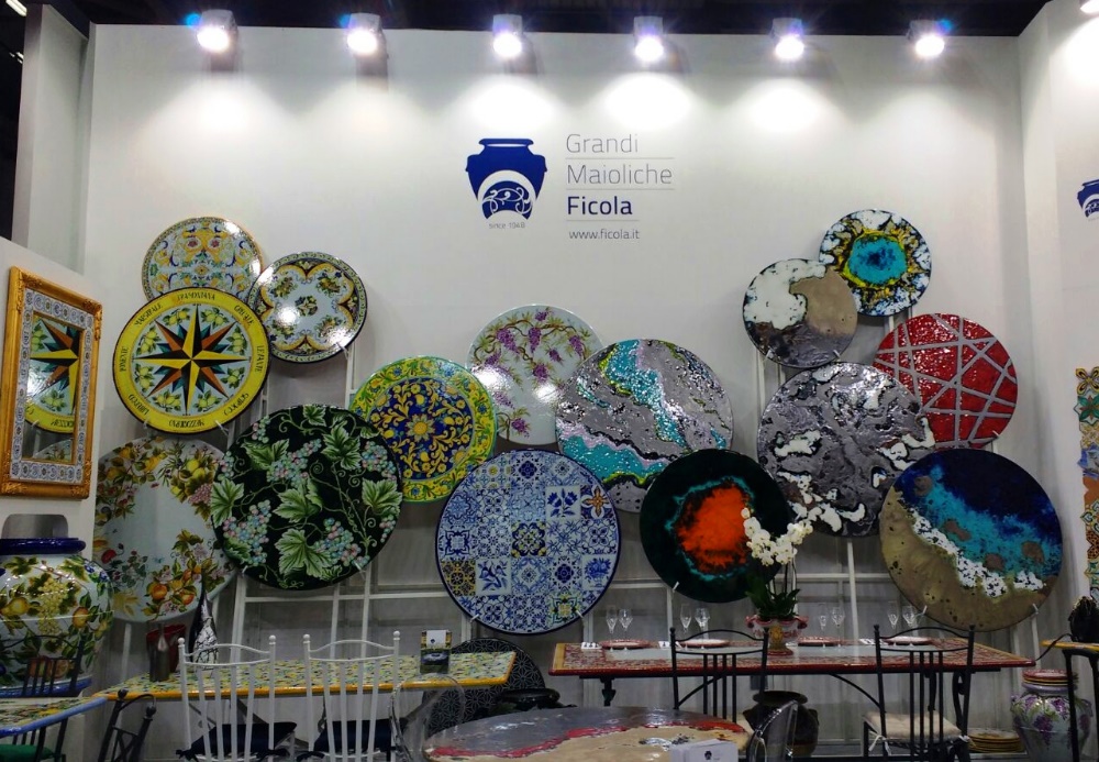 Stand espositivo "Grandi Maioliche Ficola" con piatti decorati a mano e tavoli apparecchiati.