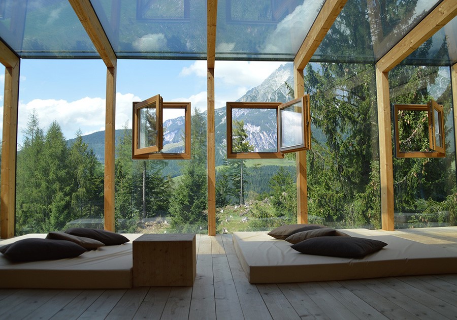 Interno moderno con tetto in vetro, finestre aperte, cuscini e vista su montagne e alberi.