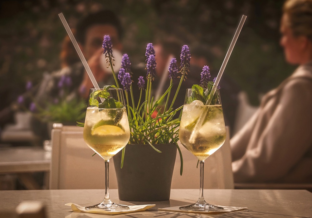 Due cocktail rinfrescanti con menta e limone su un tavolo, con fiori viola e persone sfocate sullo sfondo.