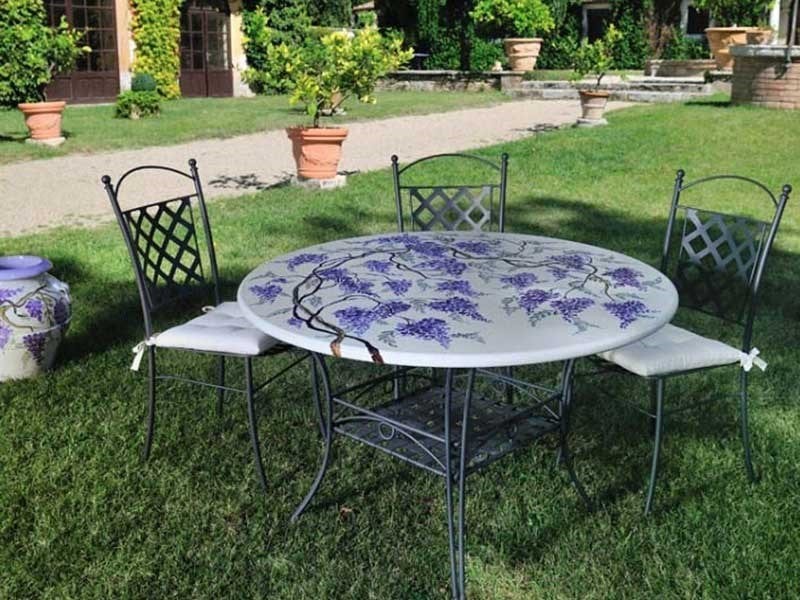 Tavolo da giardino in ferro battuto con piano decorato a glicine viola, tre sedie e cuscini bianchi su prato verde.
