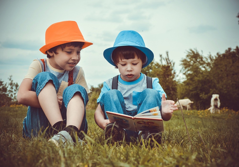 Due bambini con cappelli colorati leggono un libro seduti sull'erba.