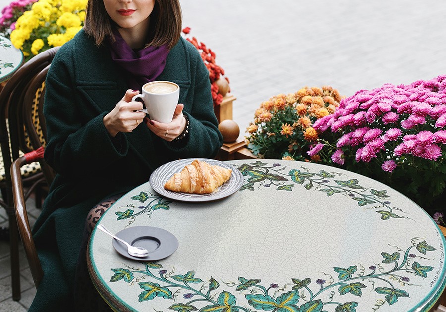 Donna con cappotto verde e sciarpa viola beve caffè al tavolo di un bar all'aperto con croissant e fiori.