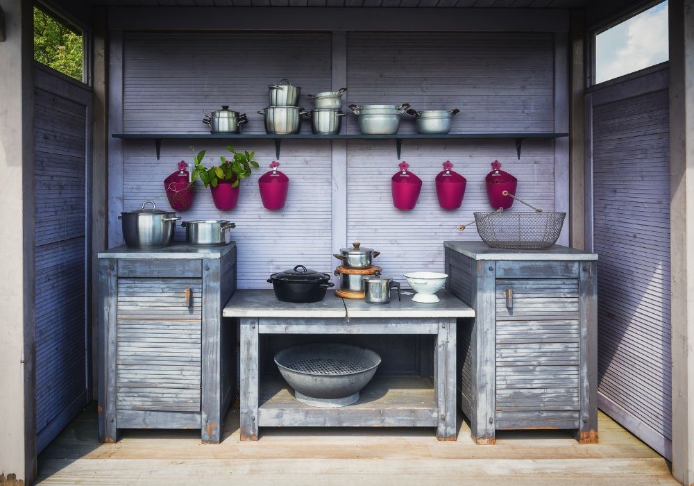 Cucina esterna in legno grigio con pentole e padelle in metallo, vasi rosa appesi e piante verdi.