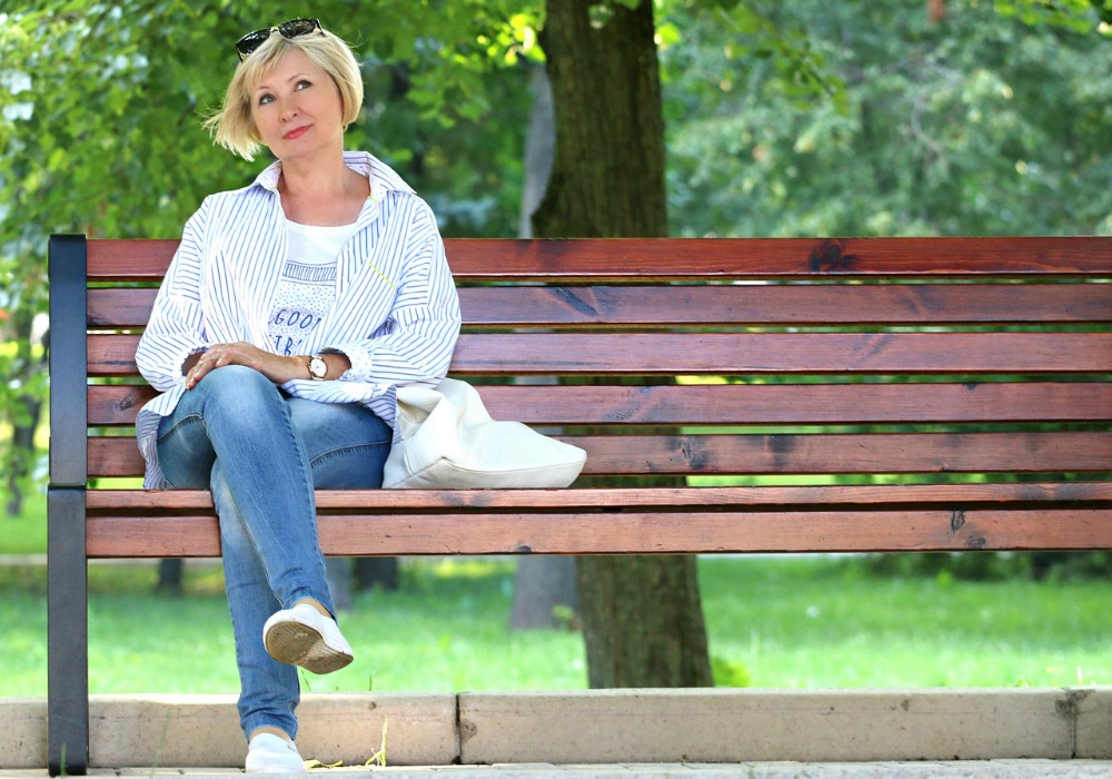 Donna bionda con camicia a righe e jeans seduta su una panchina di legno in un parco verde, borsa bianca accanto.