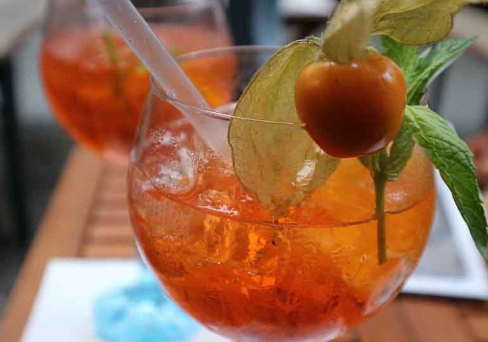Cocktail arancione con ghiaccio, cannuccia, physalis e menta, su un tavolo di legno.