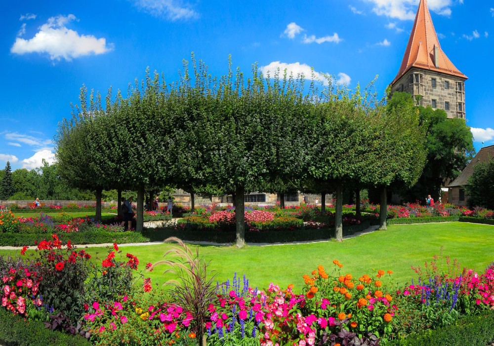 Giardino fiorito con aiuole colorate, alberi a sfera e un'antica torre in pietra sotto un cielo blu estivo.