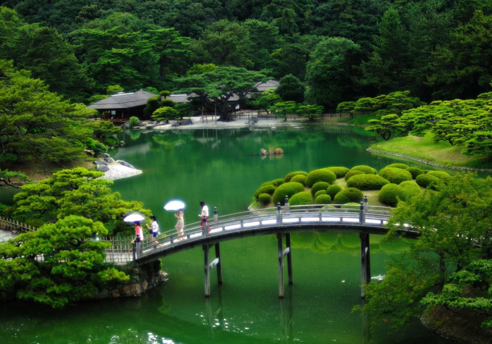 Ponte ad arco su lago verde in un giardino giapponese lussureggiante con persone e alberi.