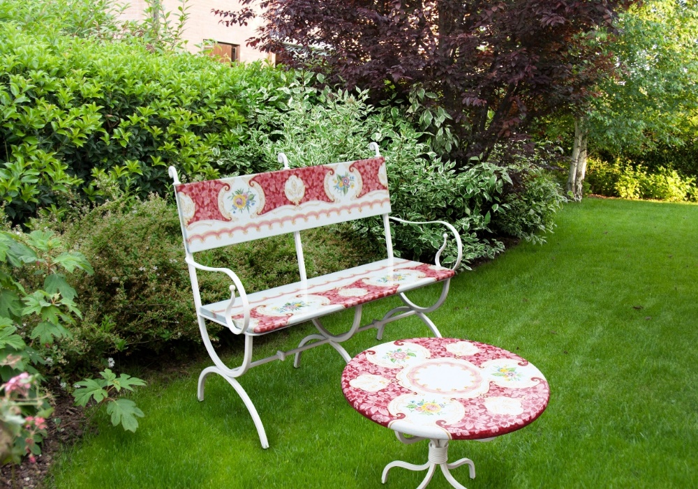 Panchina e tavolino da giardino in ferro battuto bianco con decorazioni floreali rosse in un prato verde.