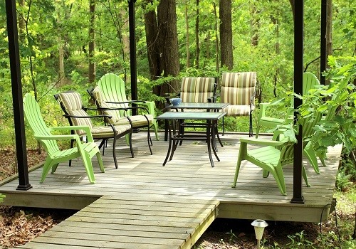 Terrazza in legno nel bosco con tavolo e sedie verdi e a strisce, sentiero in legno, alberi e vegetazione.