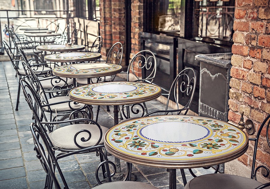 Tavoli e sedie in ferro battuto con piani in ceramica decorata all'esterno di un caffè con muro in mattoni.