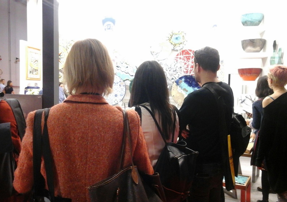 Persone osservano opere d'arte in ceramica colorate appese a una parete bianca in una galleria o fiera d'arte.