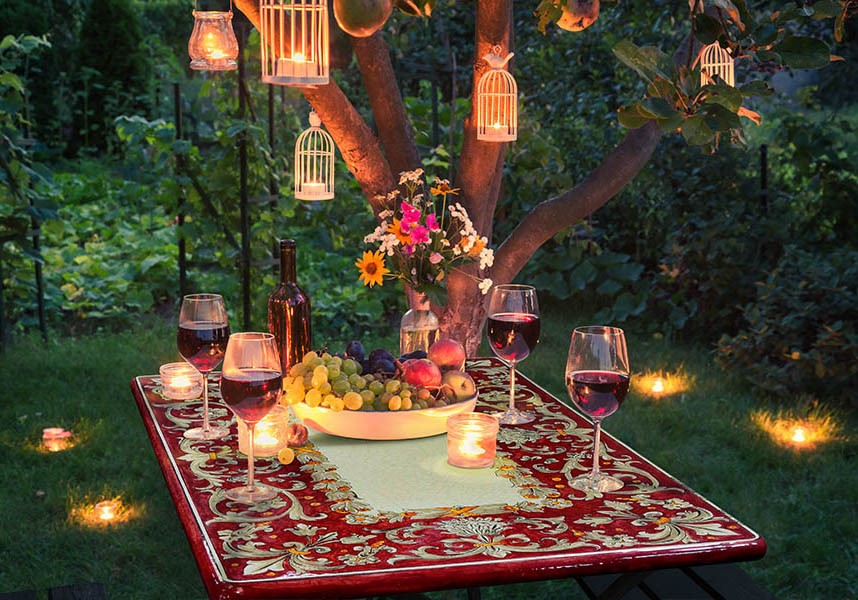 Tavolo da giardino apparecchiato per cena romantica con vino, frutta, candele e lanterne appese all'albero.