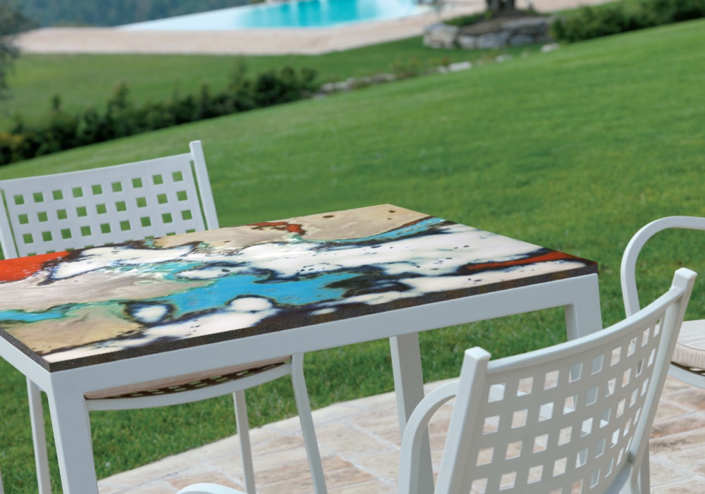 Tavolo da esterno con piano astratto colorato, sedie bianche, prato verde e piscina sullo sfondo.