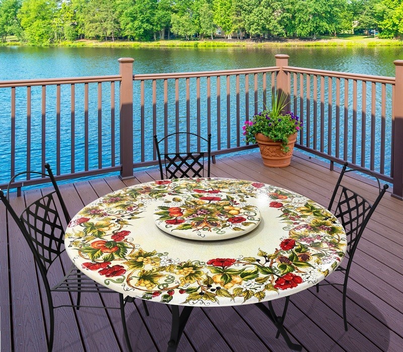 Tavolo rotondo con motivo floreale su un ponte in legno con vista lago, sedie in ferro battuto e vaso di fiori.