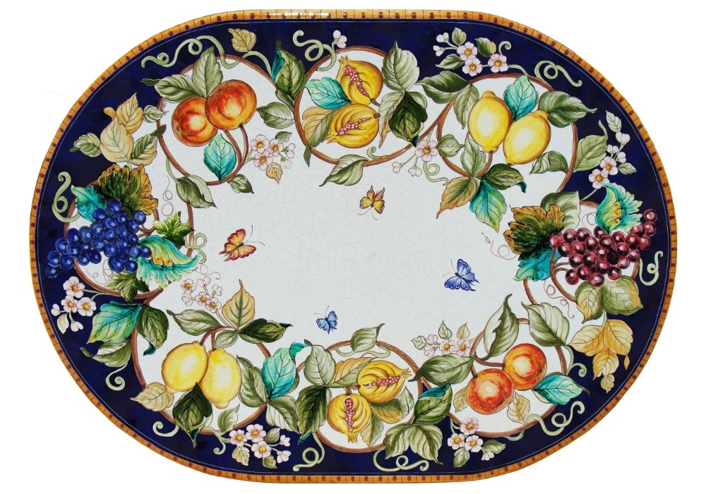 Tavolo ovale in ceramica decorato con frutta (uva, limoni, melograni, arance), foglie, fiori e farfalle su bordo blu.