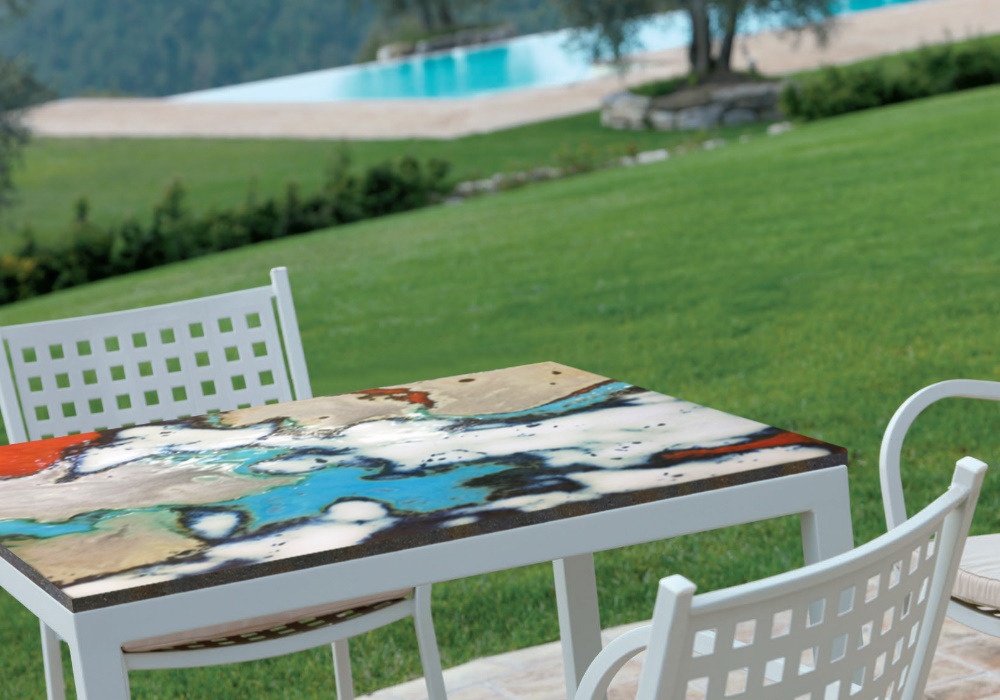 Tavolo da esterno con piano astratto colorato, sedie bianche, prato verde e piscina sullo sfondo.