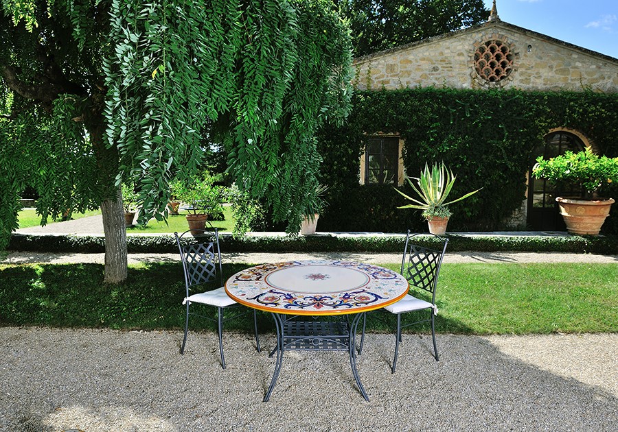 Tavolo da giardino in ceramica decorata e sedie in ferro battuto sotto un salice piangente, con villa toscana sullo sfondo.