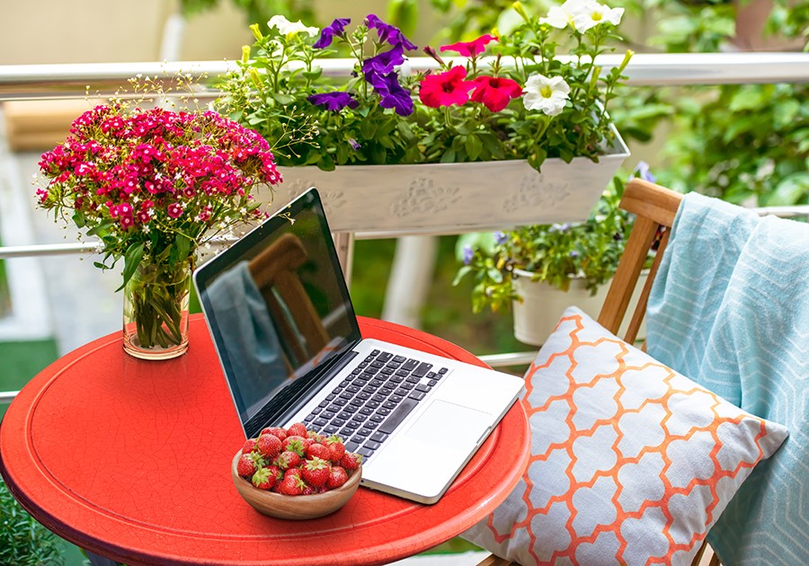 Balcone fiorito con tavolo rosso, laptop, fragole, vaso di fiori rosa e sedia con cuscino.