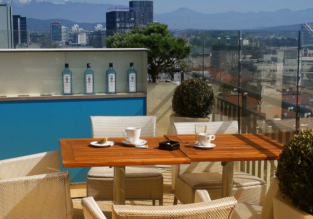 Terrazza panoramica con tavoli in legno, sedie in vimini, tazze di caffè e bottiglie di gin. Sfondo urbano e montagne.