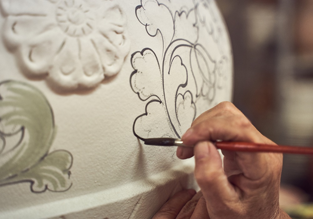 Mano che dipinge dettagli floreali neri su ceramica bianca, con motivi in rilievo e accenti verdi.