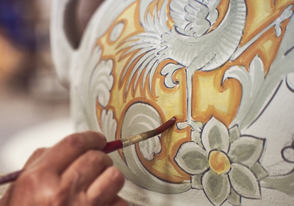 Mano dipinge motivi floreali e un uccello su ceramica bianca con colori arancio, grigio e marrone.