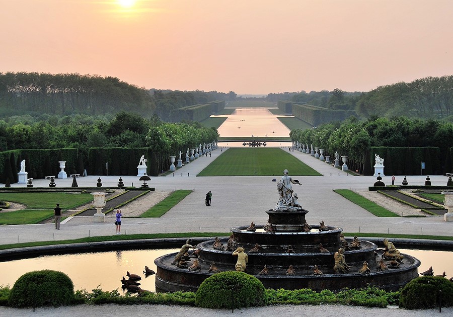 Giardini di Versailles al tramonto con fontana, statue e Grand Canal in lontananza.