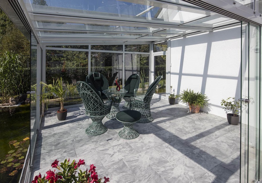 Veranda moderna con tetto in vetro, pavimento in marmo, sedie in rattan verde scuro e piante, affacciata su un laghetto.