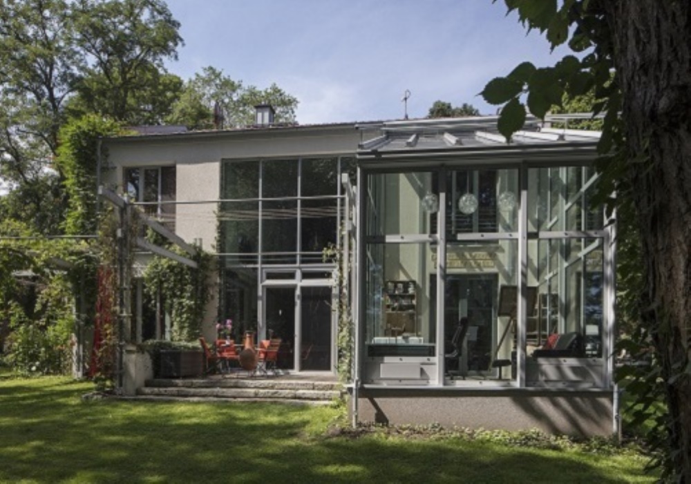 Casa moderna con ampie vetrate, giardino verde e patio esterno con tavolo e sedie rosse.